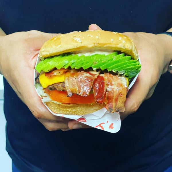Hands holding a Teriyaki J's burger with bacon, avocado, cheese, tomato, and lettuce.