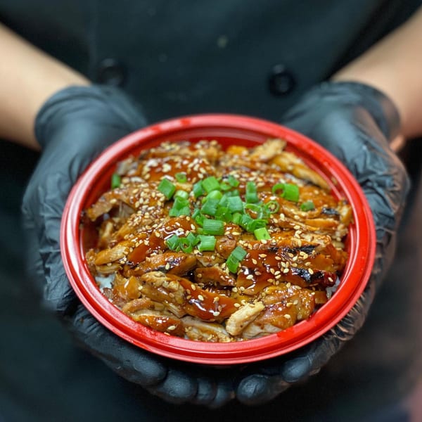 Gloved hands hold a red bowl of Teriyaki J's chicken, garnished with sesame seeds and green onions.
