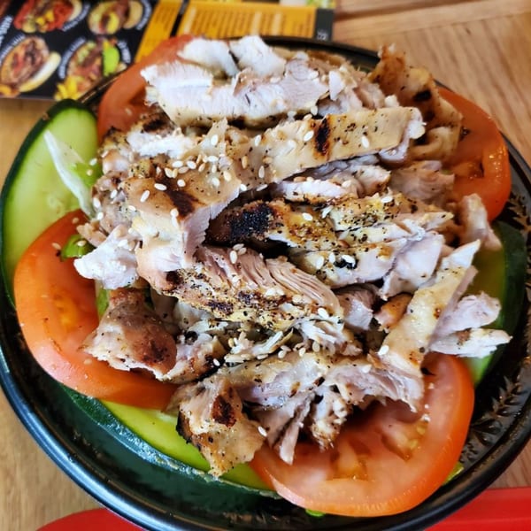Teriyaki J's grilled chicken salad with sesame seeds, tomatoes, and cucumbers in a black bowl.
